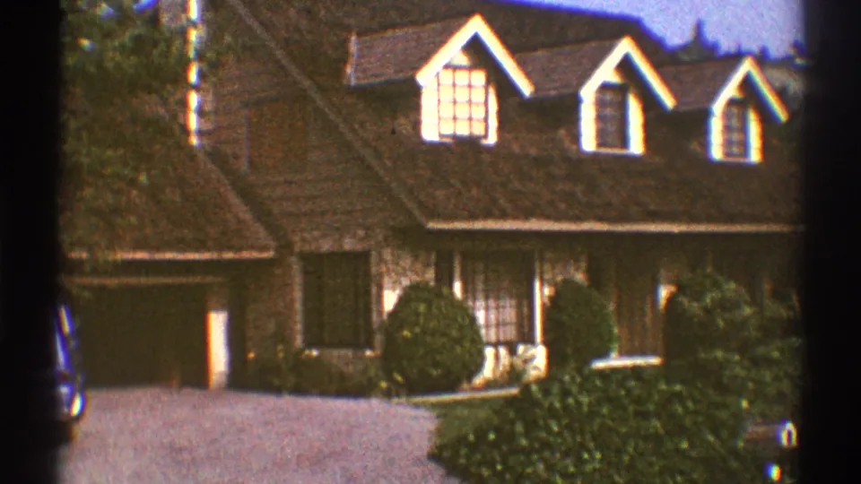 1969 Los Angeles frame with a closer home-movie view of the brown house, dormer windows, garage side, driveway, and shrubs.