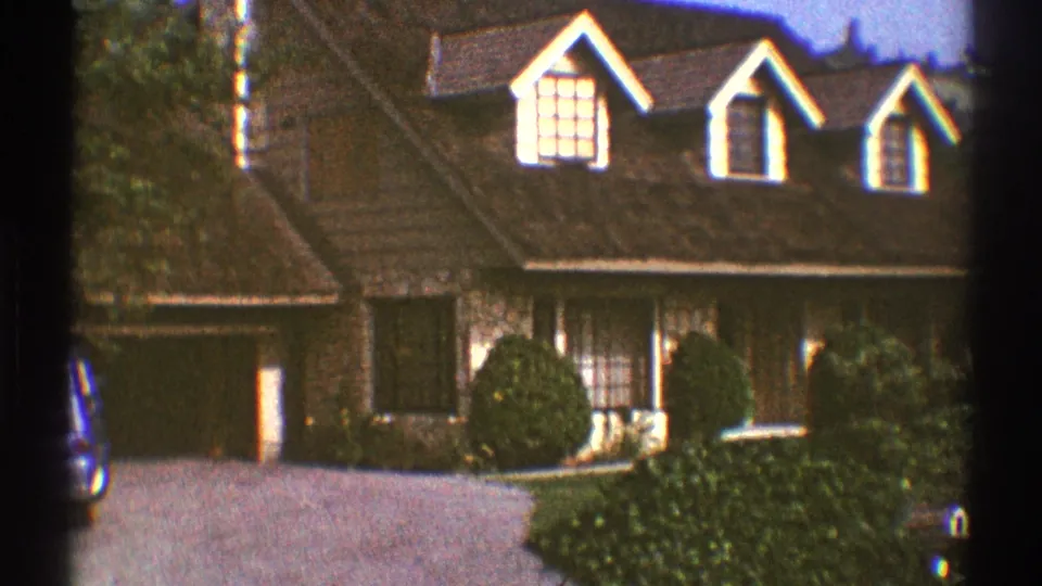 1969 Los Angeles home-movie still of a brown two-story house facade with dormer windows, shrubs, driveway, and garage at left.