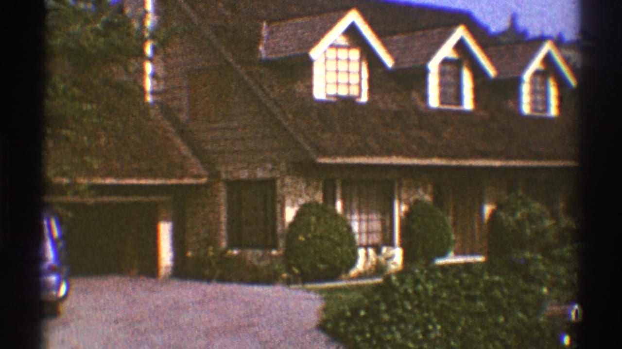 Tan suburban house with dormer windows, driveway shrubs, and part of a parked car at the frame edge.