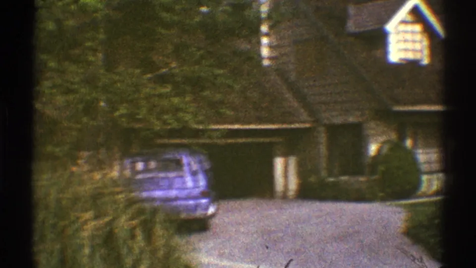 1969 Los Angeles suburban scene with a blue car parked by an open garage at a tan house with trees and shrubs around the driveway.