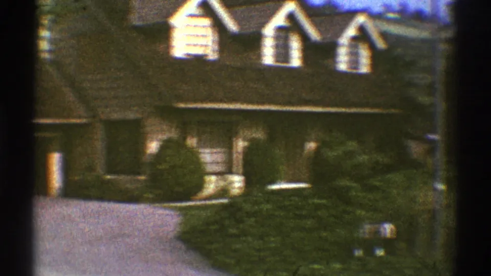 1969 Los Angeles home-movie frame with a tan residential facade, dormer windows, dense shrubs, driveway, and partial car at the edge.