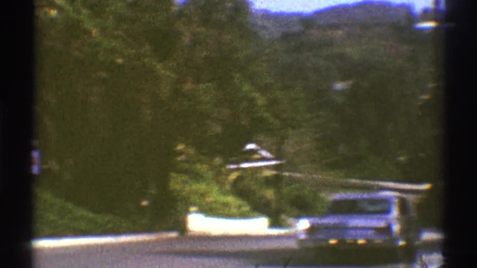 1969 Los Angeles street view from a moving car, with a light sedan, white curb, greenery, and hillside background.