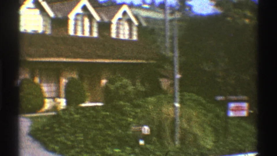 1969 Los Angeles residential exterior with gabled windows, dense hedges, a sidewalk, and a small for-sale sign.