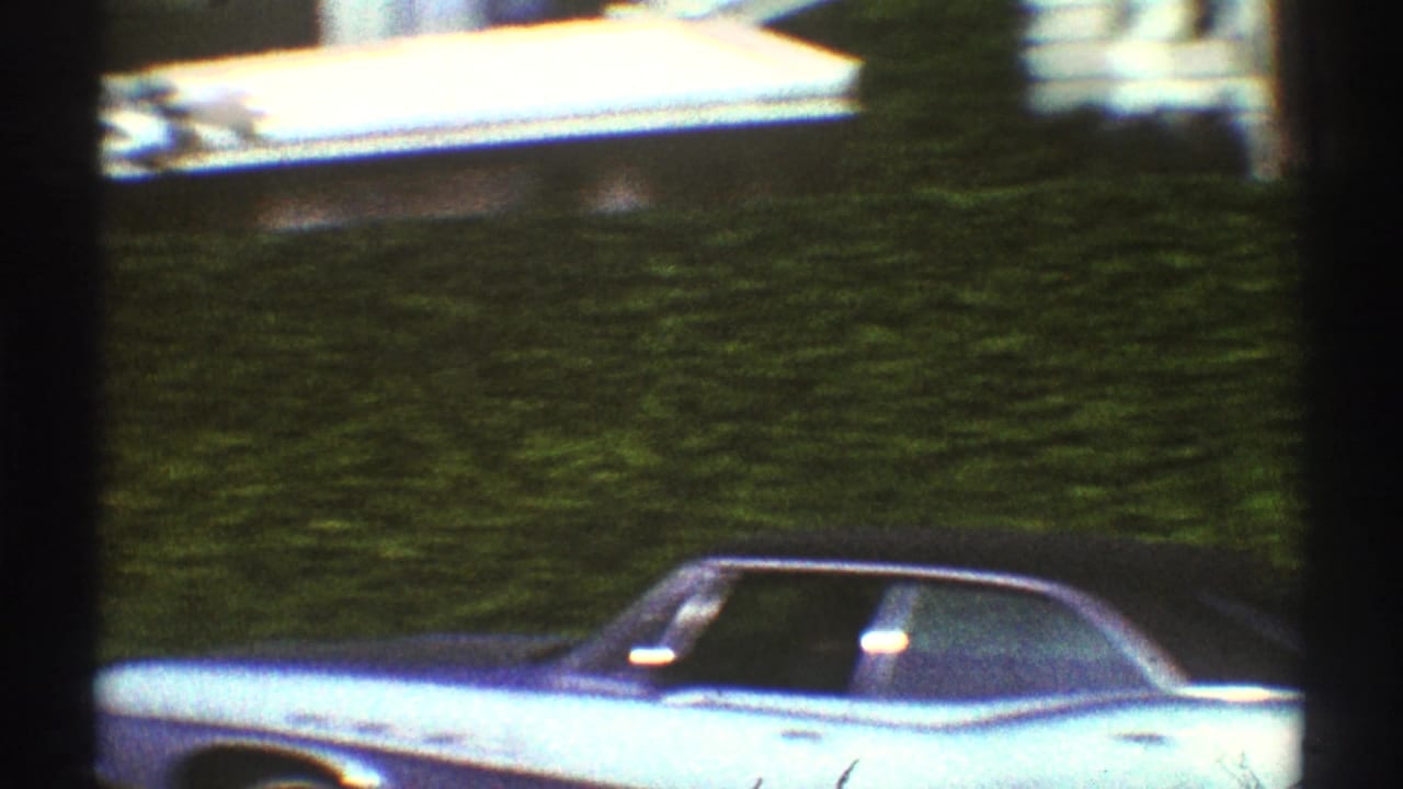 1969 Los Angeles home-movie frame showing a light blue car parked along a curb beside dense green hedges.