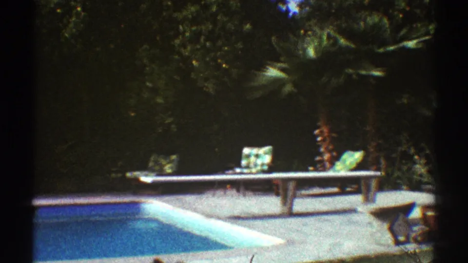 1969 Los Angeles 8mm poolside view with blue water, lounge chairs, patio paving, a palm tree, and dense garden foliage.