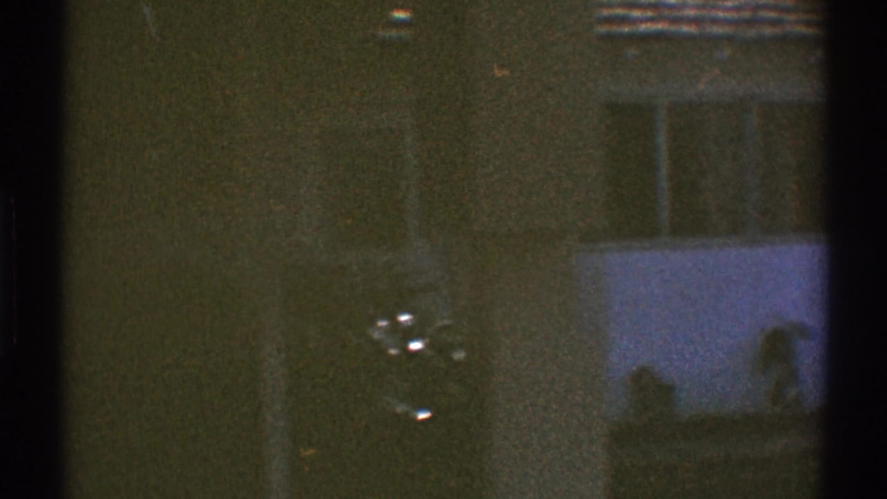 Grainy 1969 Los Angeles home-movie frame showing part of a white residential exterior with windows seen through dark foreground framing.