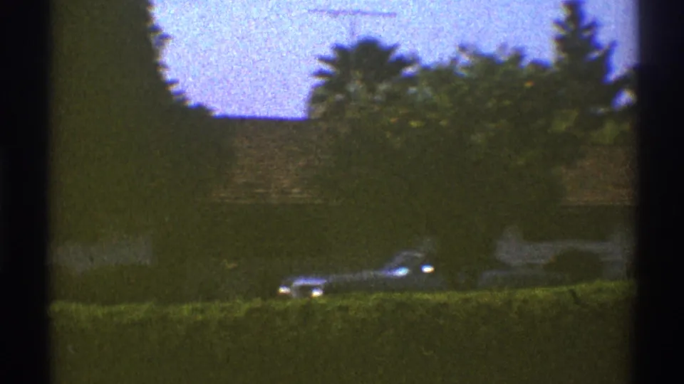 1969 Los Angeles home-movie view across a hedge toward a masonry wall and a partially hidden parked car.