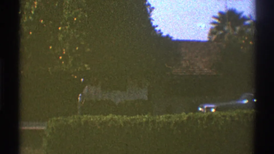 1969 Los Angeles 8mm still showing a hedge-lined exterior with a partly obscured classic car and masonry wall.