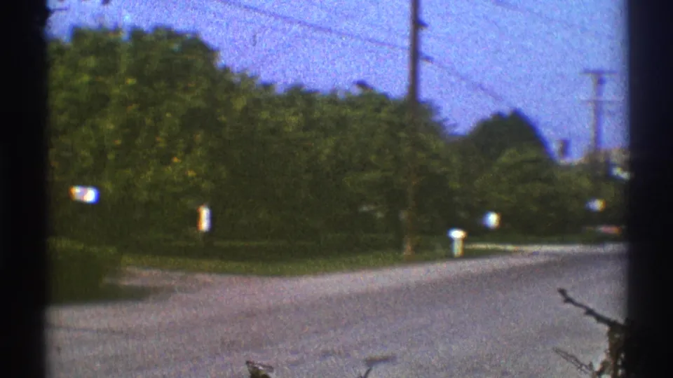 1969 Los Angeles suburban road with mailboxes, overhead utility lines, trees, and vehicles seen from a home-movie perspective.