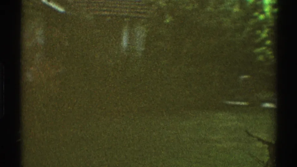 1969 Los Angeles home-movie frame showing a shaded driveway, partially obscured parked car, grass, foliage, and a dark circular stain on concrete.