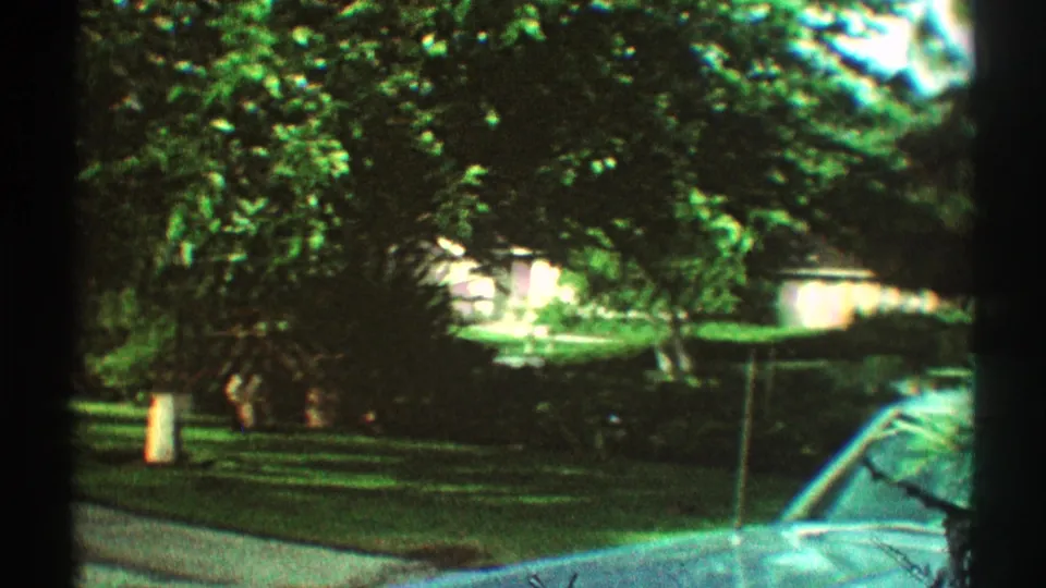 1969 Los Angeles frame with a blue parked car at right, a curving residential street, green lawn, and dense trees around a partially hidden house.