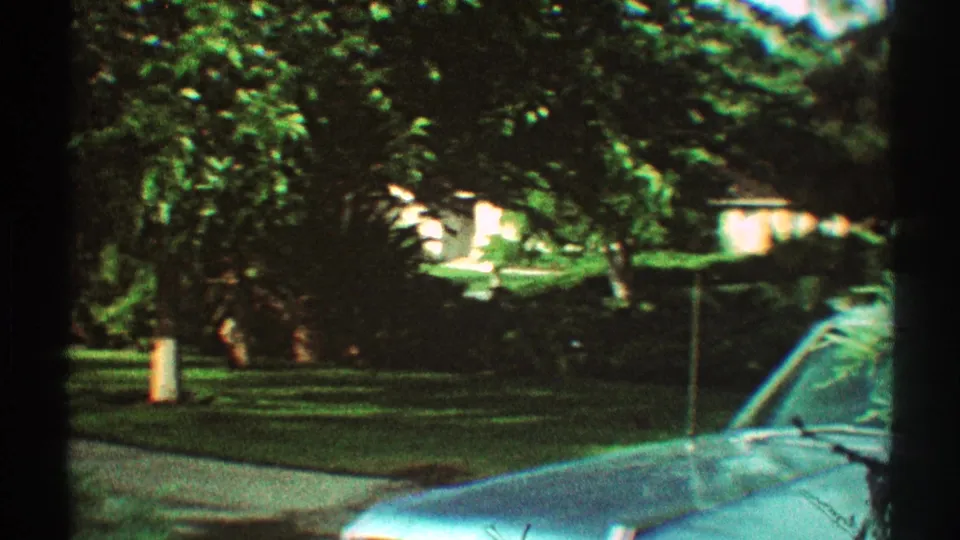 1969 Los Angeles home-movie view of a blue parked car beside a shaded residential lawn, with trees and a house partially visible through foliage.