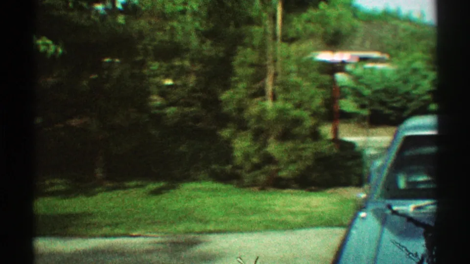 1969 Los Angeles home-movie frame showing a parked car edge, green lawn, trees, a utility pole, and roadside landscaping.