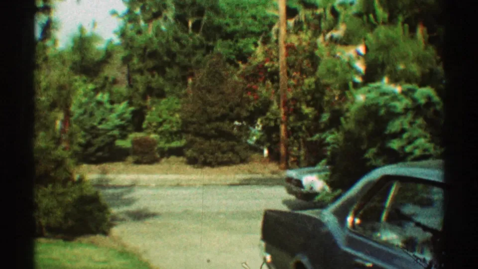 1969 Los Angeles home-movie frame with a blue car in the foreground, curved driveway, green lawn, shrubs, and trees along a quiet street.