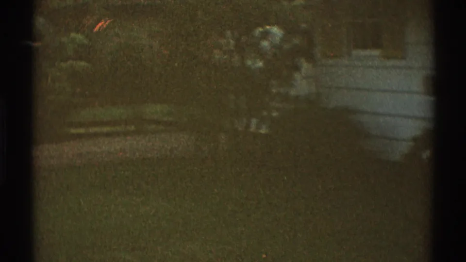 1969 Los Angeles home-movie frame showing a shadowed grassy area with shrubs, trees, and a pale structure in the background.
