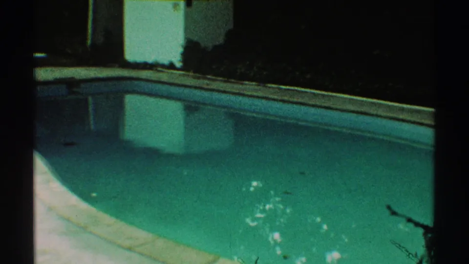 1969 Los Angeles home-movie frame showing a backyard pool at night, with turquoise water, pale coping, and dark surrounding foliage.