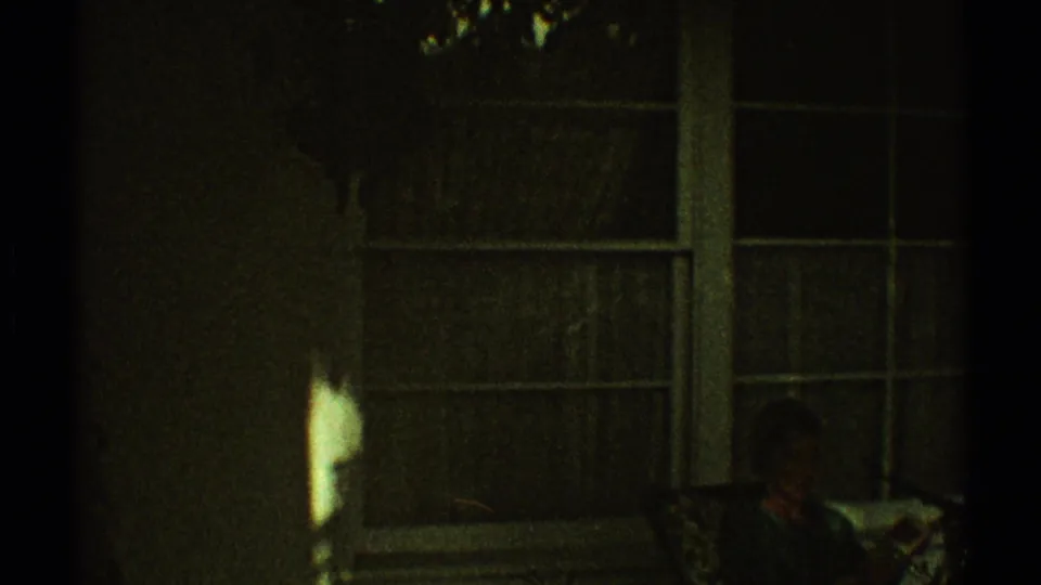 1969 Los Angeles frame looking toward a dark windowed interior, where a faint seated person appears near the lower right.