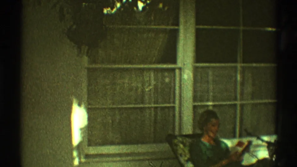 In 1969 Los Angeles home-movie footage, a woman sits on a porch chair reading near a large curtained window in soft light.