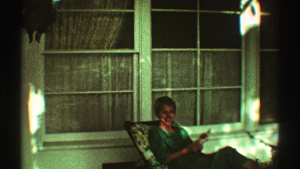 A woman in a green dress smiles while reading on a porch chair in 1969 Los Angeles home-movie footage.