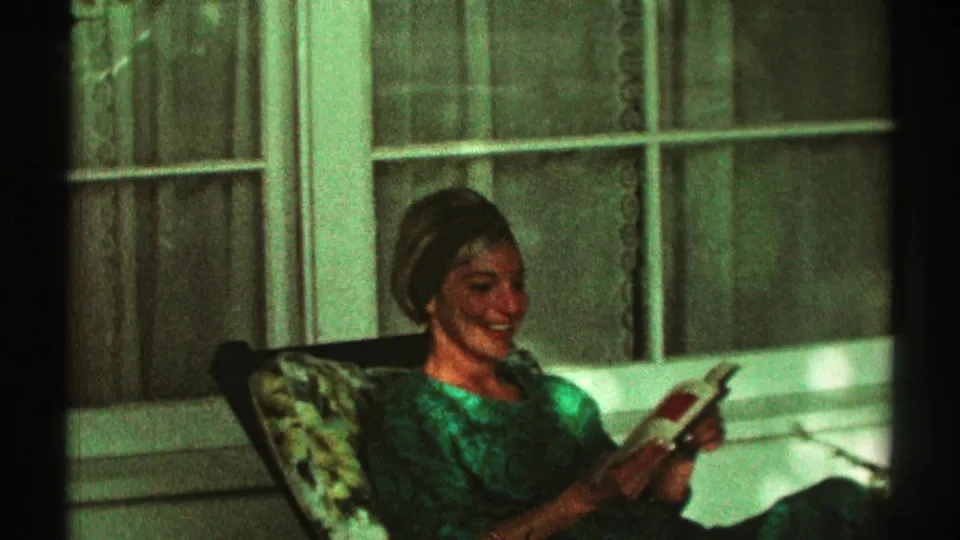 Closer 1969 Los Angeles frame of a woman smiling as she reads a book in a porch chair beside a curtained window.