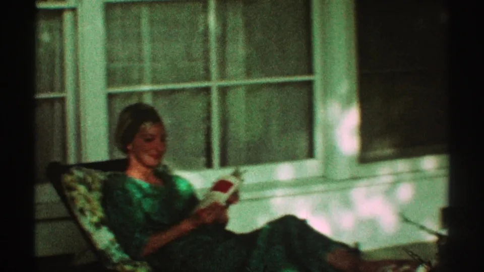 A woman reclines in a porch chair with an open book near a large window in 1969 Los Angeles home-movie footage.