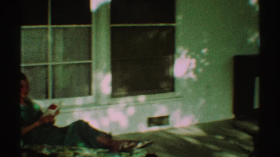 In 1969 Los Angeles home-movie footage, a woman in a green dress sits on a porch chair reading near windows and dappled shade.
