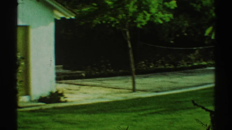 1969 Los Angeles residential yard frame showing a white house exterior, paved area, chain-link fence, lawn, and small tree.
