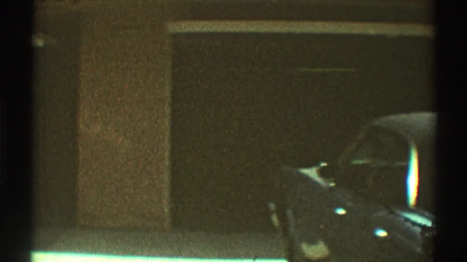 1969 Los Angeles exterior frame showing a shaded house wall, carport or driveway opening, and part of a parked dark car.