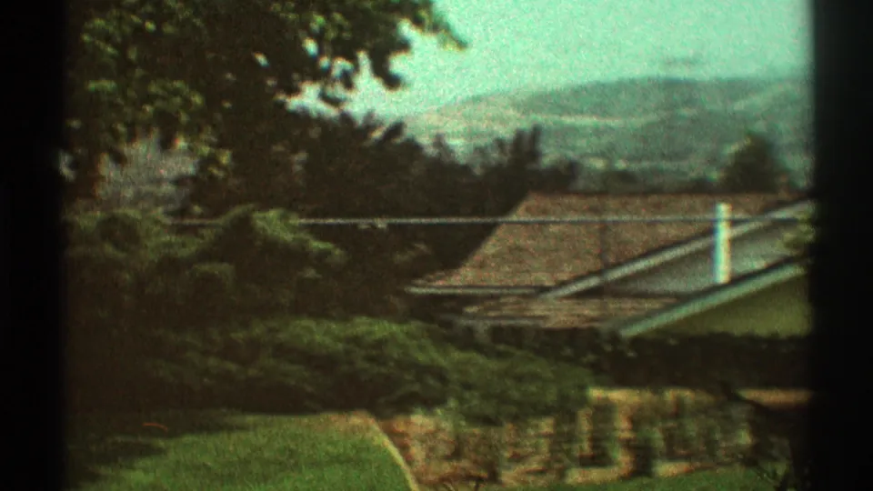 1969 Los Angeles residential view with rooflines, garden hedges, trees, and distant hills in soft 8mm color.