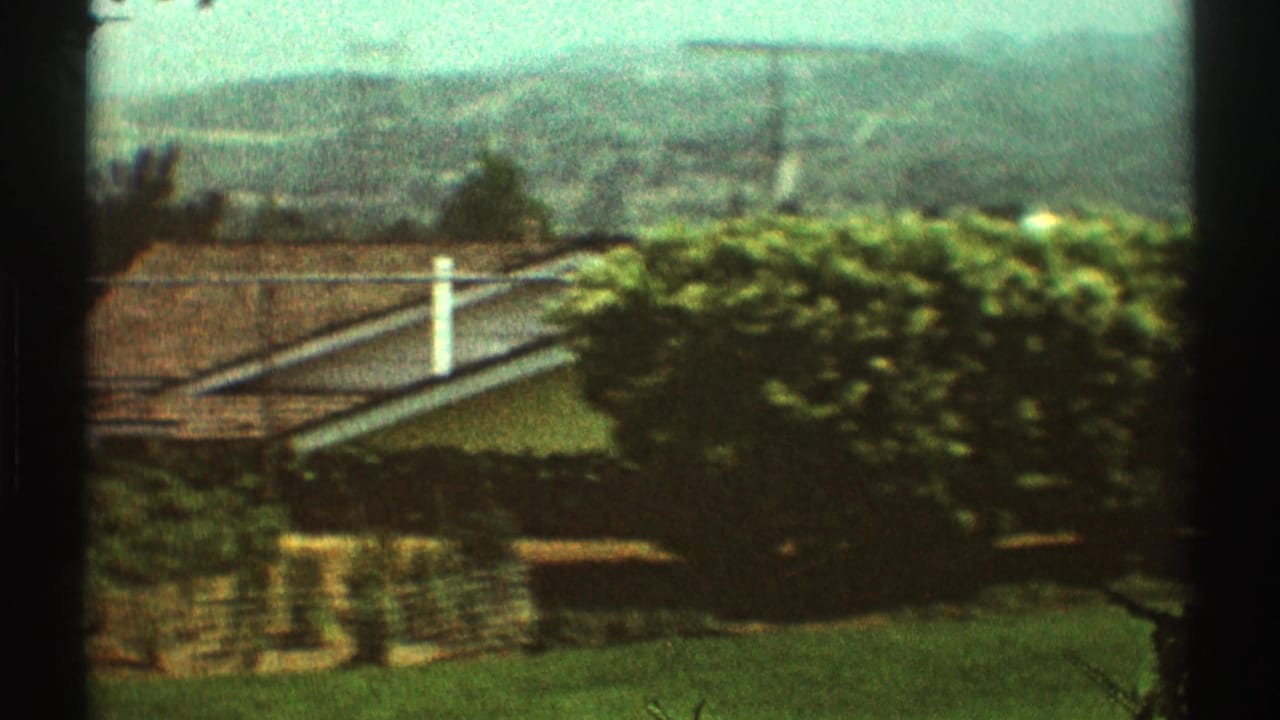 1969 Los Angeles home-movie view across a backyard lawn toward hedges, a roofline, chimney, and hazy hills.