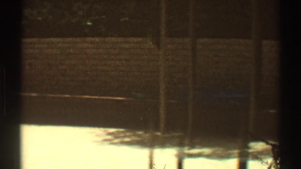 1969 Los Angeles home-movie frame showing a patio or walkway edge, wood fencing, and a heavily shaded yard area.