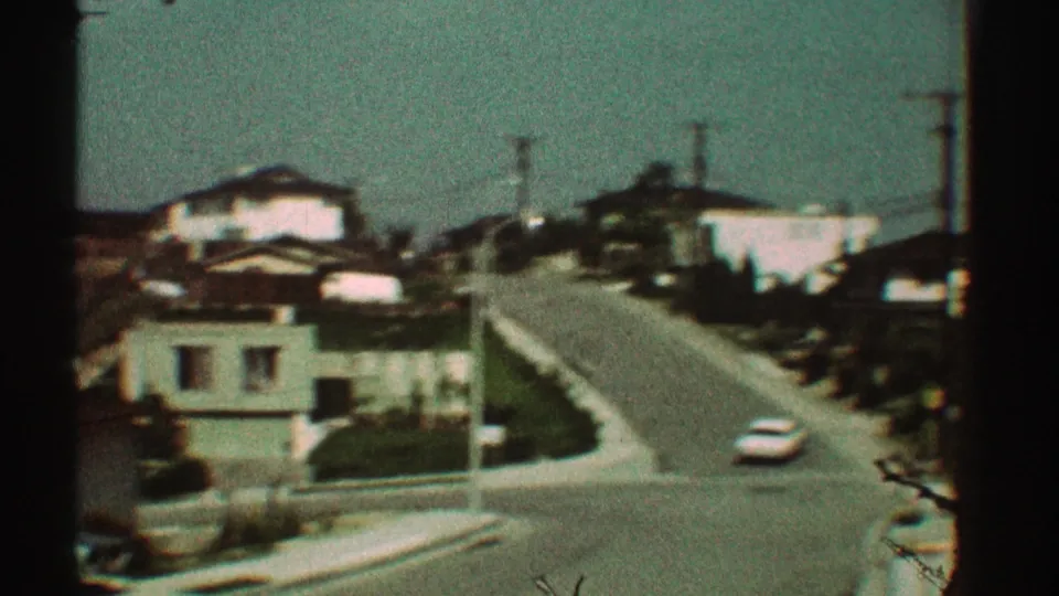 1969 Los Angeles home-movie view of a white car driving uphill past low residential buildings and utility poles.