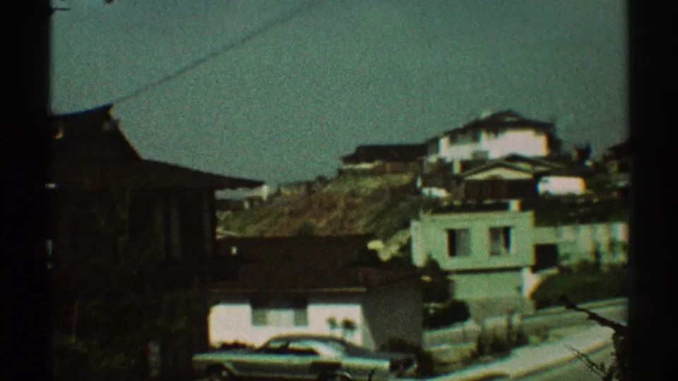 1969 Los Angeles frame showing hillside homes, a parked car, shrubs, and an overhead wire in soft 8mm color.