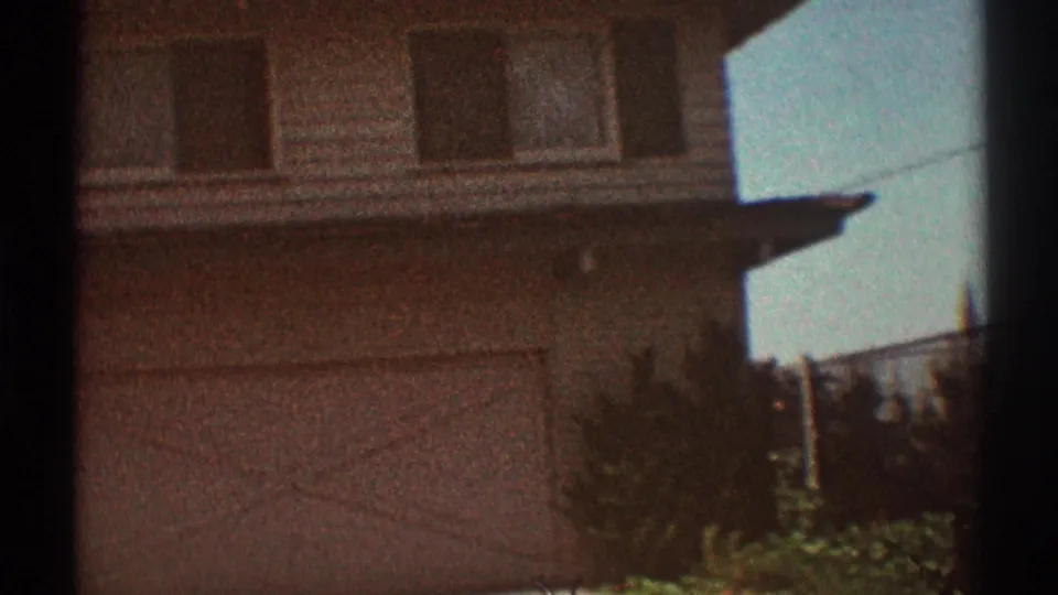 1969 Los Angeles home-movie frame with a residential facade, upper windows, roofline, shrubs, and fence at the edge of the shot.