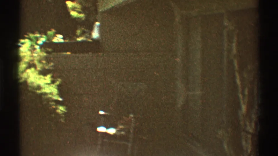 1969 Los Angeles 8mm frame with a brick wall, metal patio chair, shaded exterior area, and sunlit garden plants.