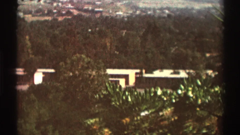 1969 Los Angeles home-movie frame showing a distant residential exterior beyond leafy garden plantings, with soft focus and dark frame edges.