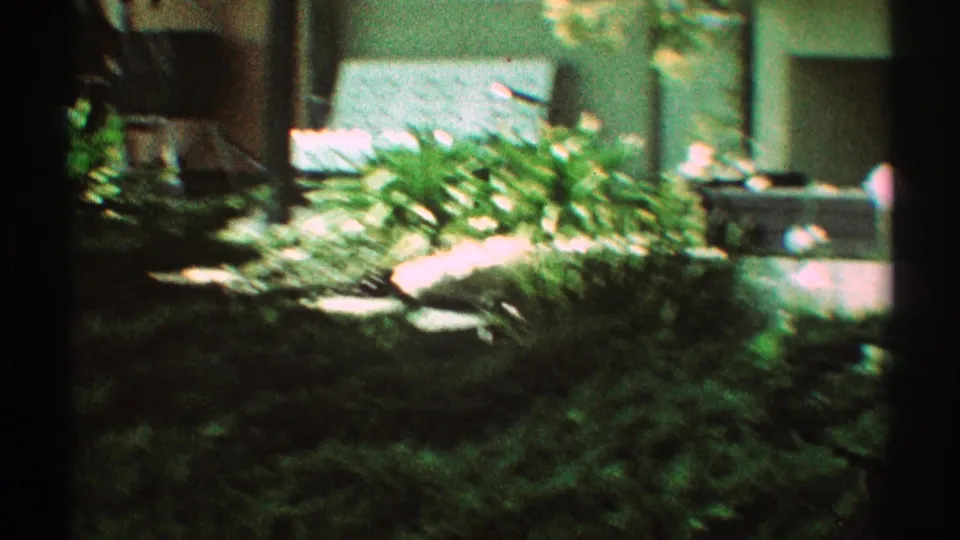 Home-movie frame of a Los Angeles residential garden, showing shrubs, sunlit paving, shaded plantings, and partial house exterior detail.