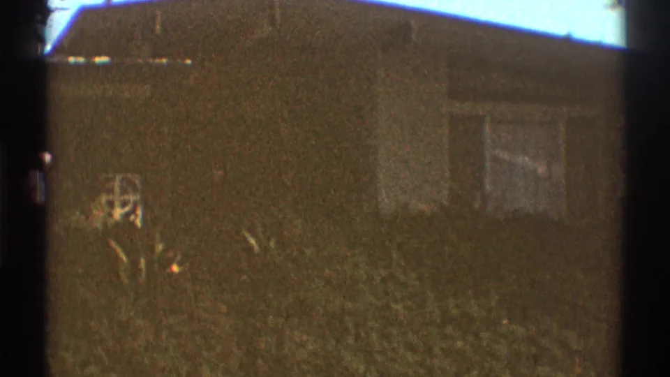 1969 Los Angeles home-movie frame of a residential house exterior with windows, lawn, shrubs, roofline, and washed bright sky.