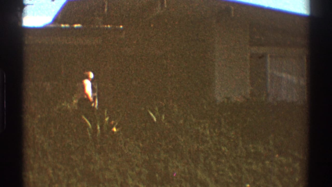 In 1969 Los Angeles home-movie footage, a person in a white shirt crosses a grassy yard near a low residential building.