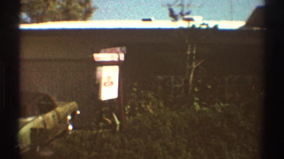 A 1969 Los Angeles home-movie frame shows a driveway edge, partial vintage car, house exterior, vegetation, and a small sign.