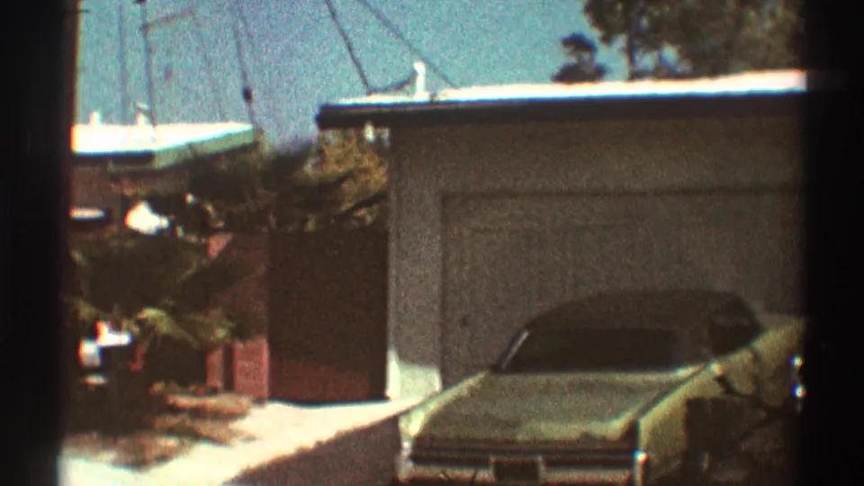 1969 Los Angeles home-movie frame showing a parked car in a driveway beside a garage and low residential buildings.