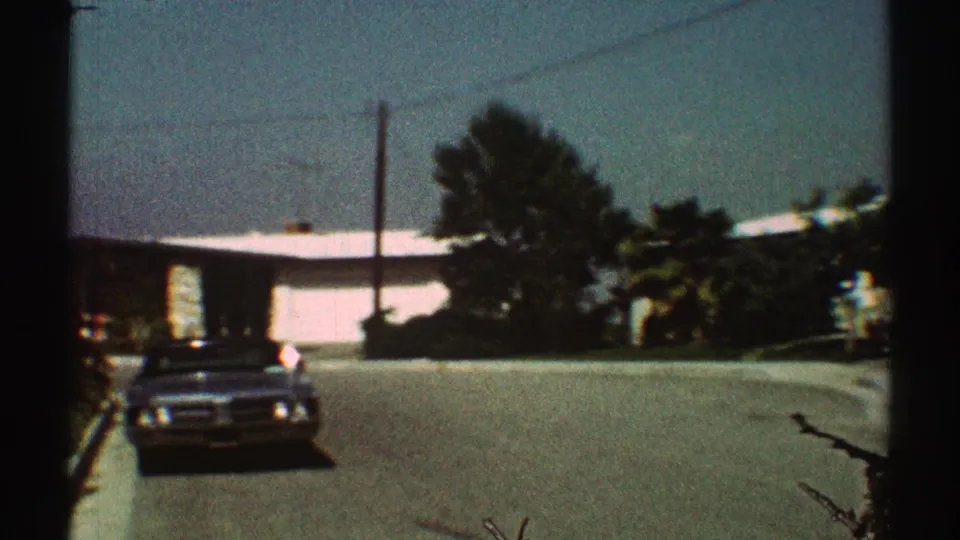 1969 Los Angeles home-movie frame showing a parked car, a distant person, trees, and single-story homes on a sunny street.