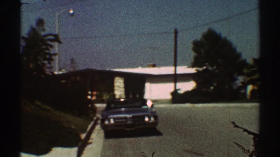 1969 Los Angeles home-movie frame showing a dark sedan on a sunlit residential street lined with low houses and palm trees.