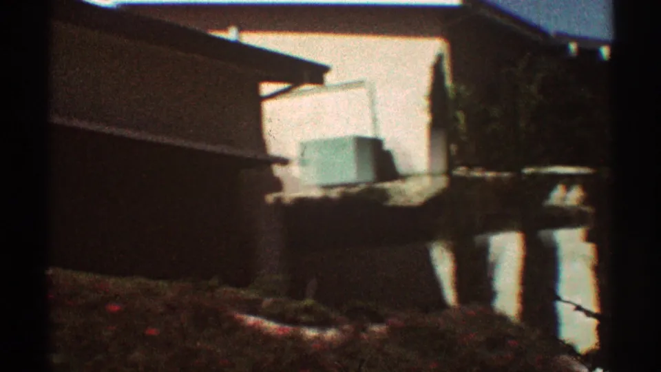 1969 Los Angeles home-movie frame showing a residential side yard, stucco wall, small outbuilding or enclosure, and trees.