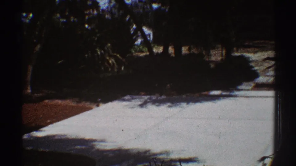 End of the backyard pan showing a bright concrete patio, heavy tree shade, shrubs, and fencing in grainy 8mm footage.