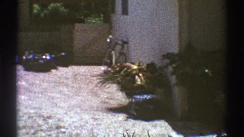 1969 Los Angeles home-movie frame with a bicycle by a wall, potted plants, and scattered backyard objects.