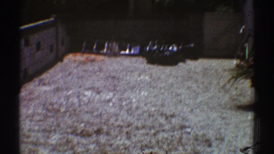 Late-1960s 8mm frame of a residential yard with a bright ground surface, wall, plants, and a ladder or rail nearby.