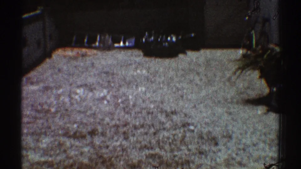 The camera remains low on the gravel driveway, with dark film borders and a shaded structure just entering view.