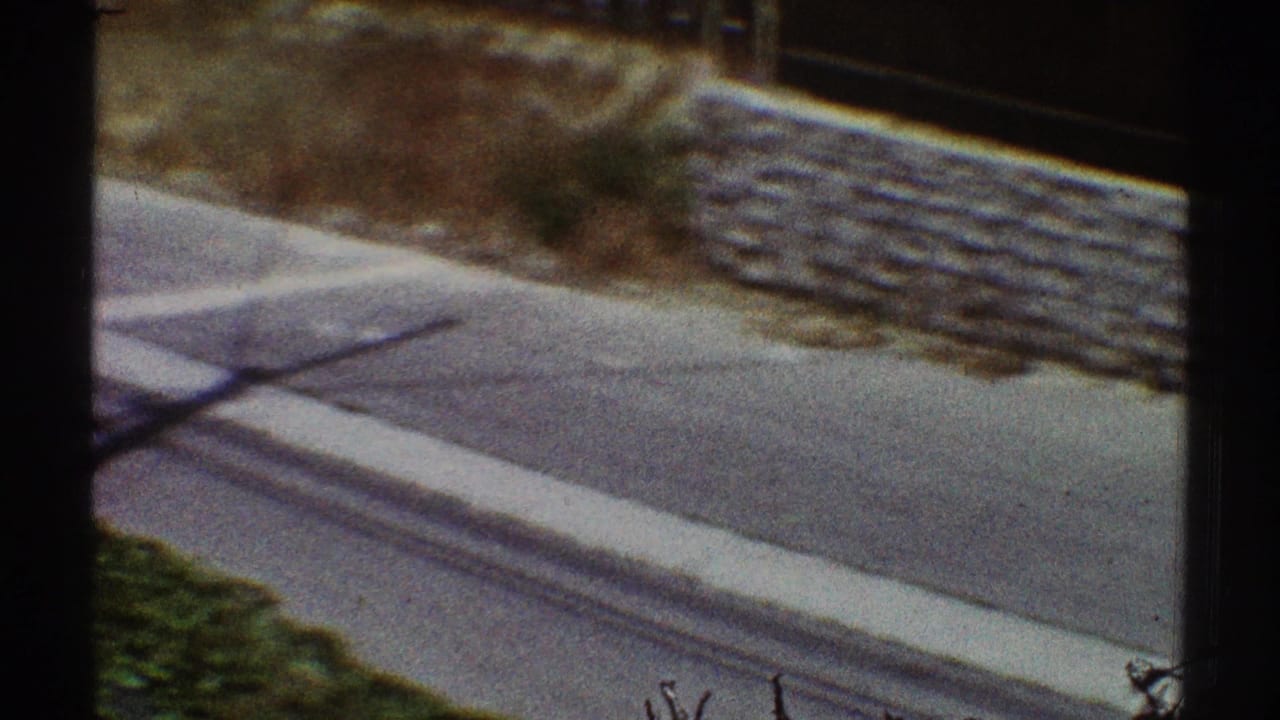 1969 Los Angeles 8mm frame looking down toward a sidewalk, street edge, stone wall, and a long shadow across the pavement.