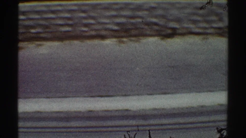 1969 Los Angeles 8mm frame with asphalt, curb, and a stacked stone wall seen from an elevated position through dark foreground framing.